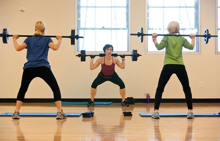Women following the instructor in doing back squats with weighted barbells.
